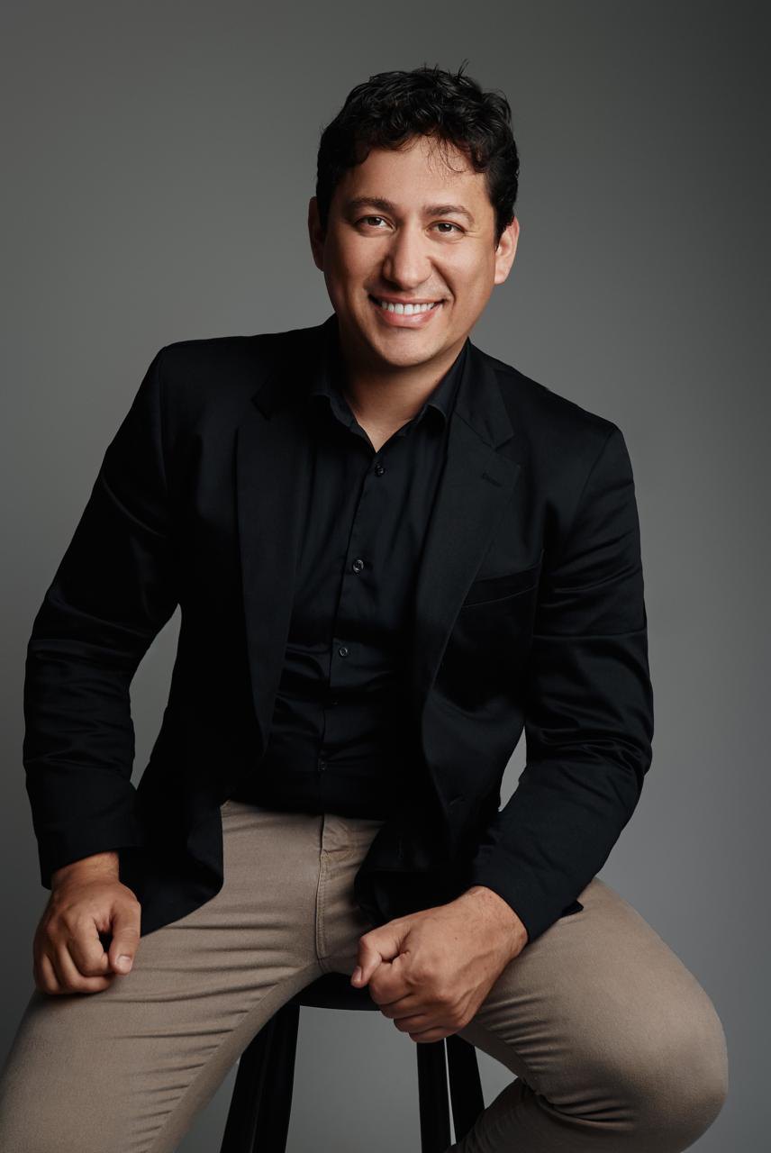 Dr. Rodrigo Campanholo sorrindo, de blazer preto, sentado em um banco